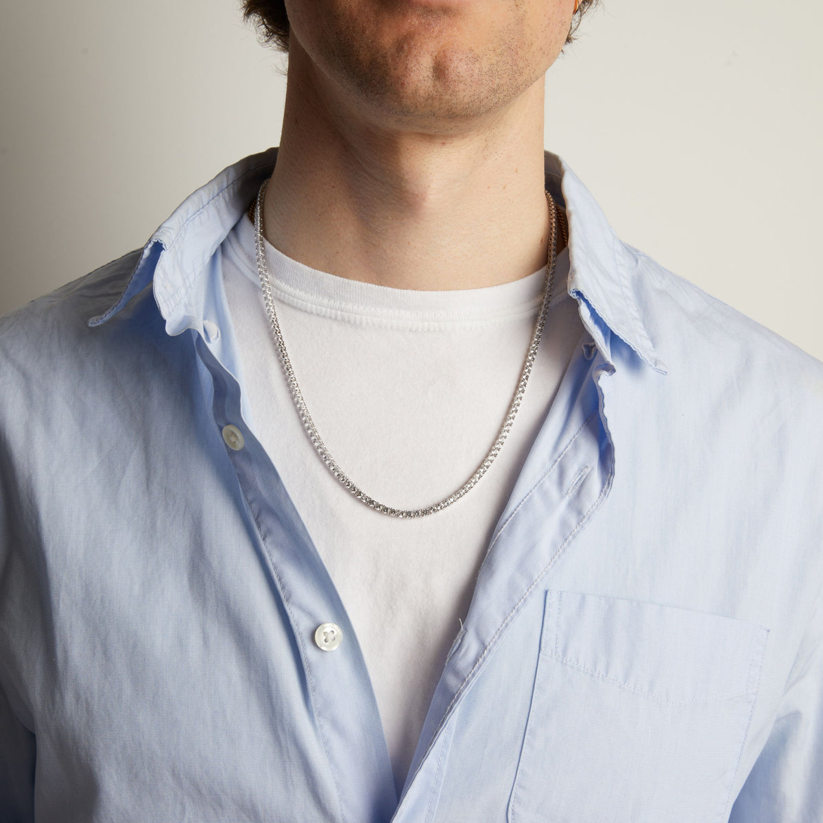 A close-up of a man wearing a tennis necklace over a white t-shirt and a light blue button-down shirt.
