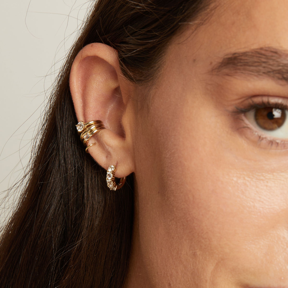 Model. Close up of woman wearing three gold ear cuffs and a huggie earring with white sapphires on neutral background. 