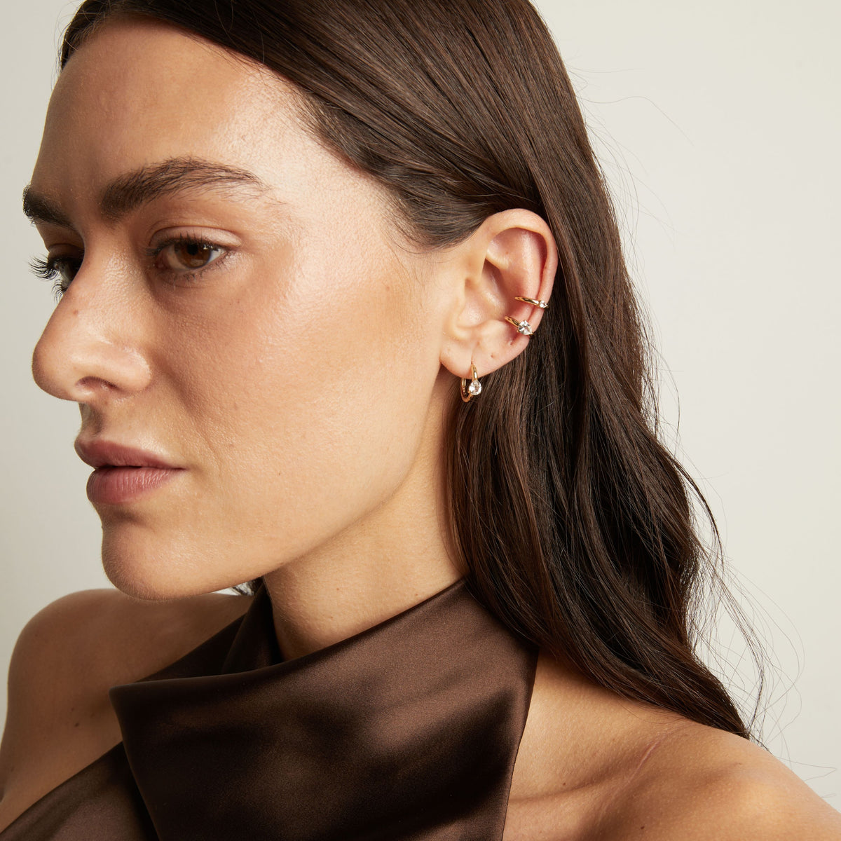 Model. Close up of woman wearing two gold ear cuffs and a huggie earring with white sapphires on neutral background. 