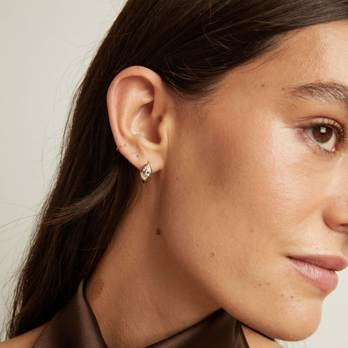 Model. Close up of woman wearing a gold bezel huggie earring on neutral background.