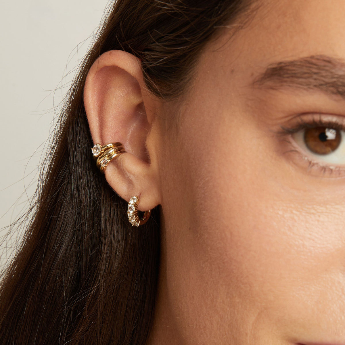 Model. Close up of woman wearing three gold ear cuffs and a huggie earring with white sapphires on neutral background. 