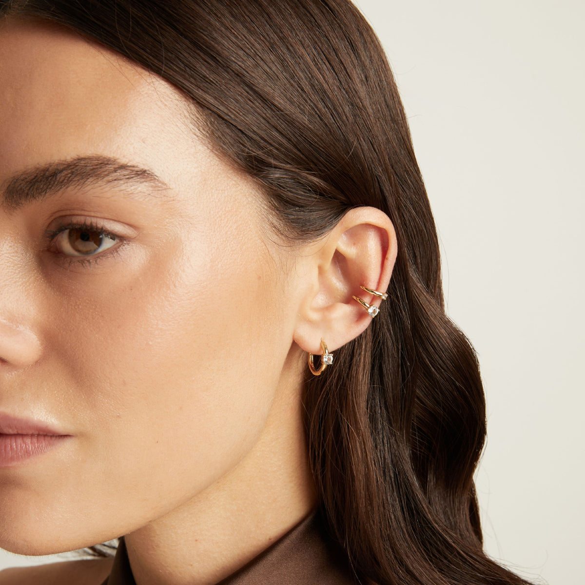 Model. Close up of woman wearing two gold ear cuffs and a huggie earring with white sapphires on neutral background. 