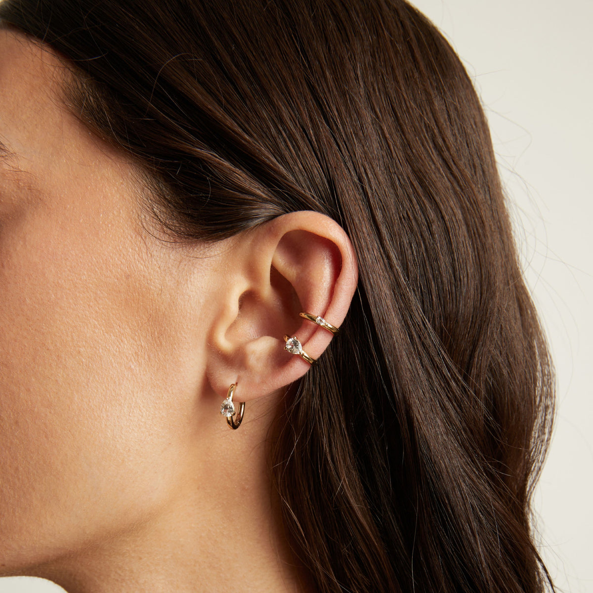Model. Close up of woman wearing two gold ear cuffs and a huggie earring with white sapphires on neutral background. 