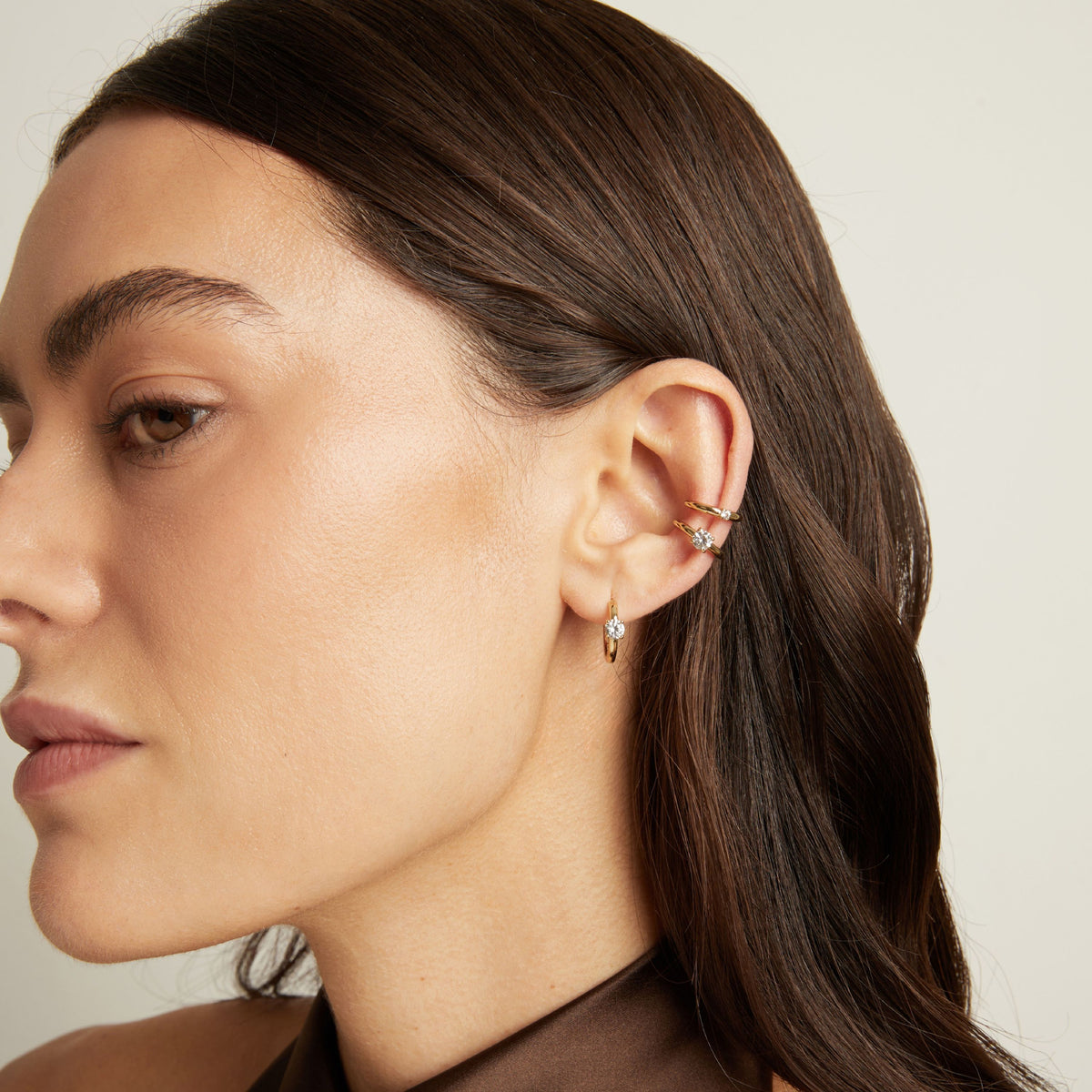 Model. Close up of woman wearing two gold ear cuffs and a huggie earring with white sapphires on neutral background. 