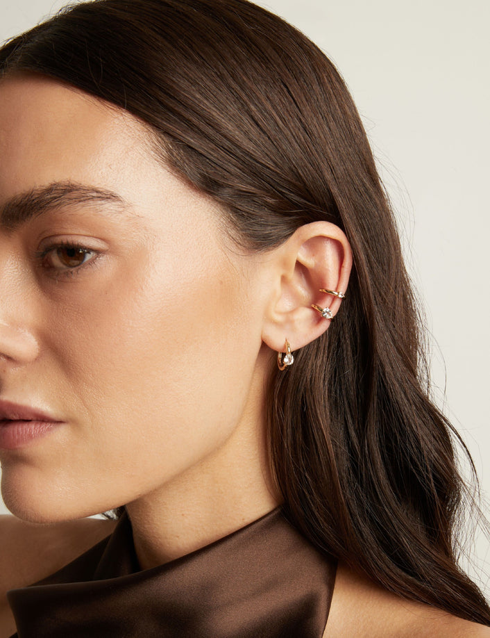 Close up of woman wearing two gold ear cuffs and a huggie earring with white sapphires on neutral background. 