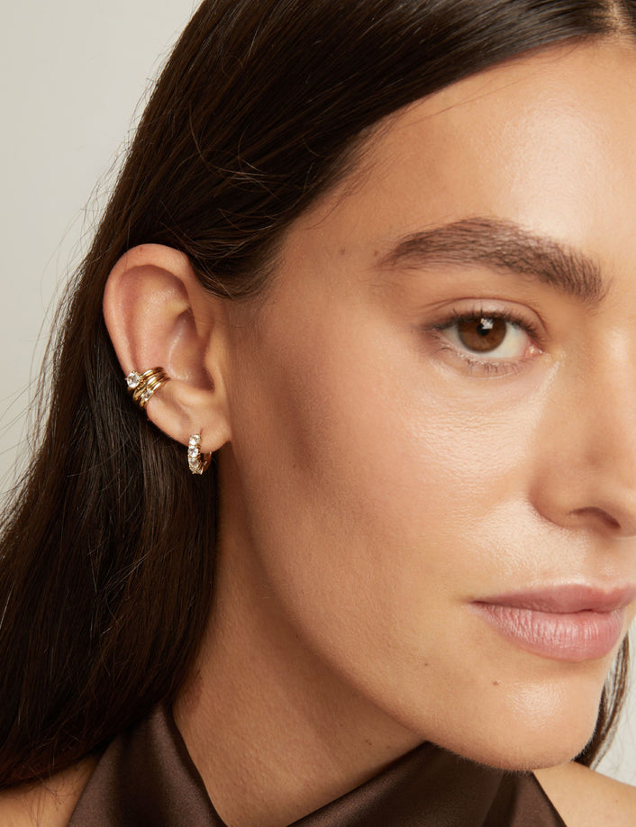 Model. Close up of woman wearing three gold ear cuffs and a huggie earring with white sapphires on neutral background. 