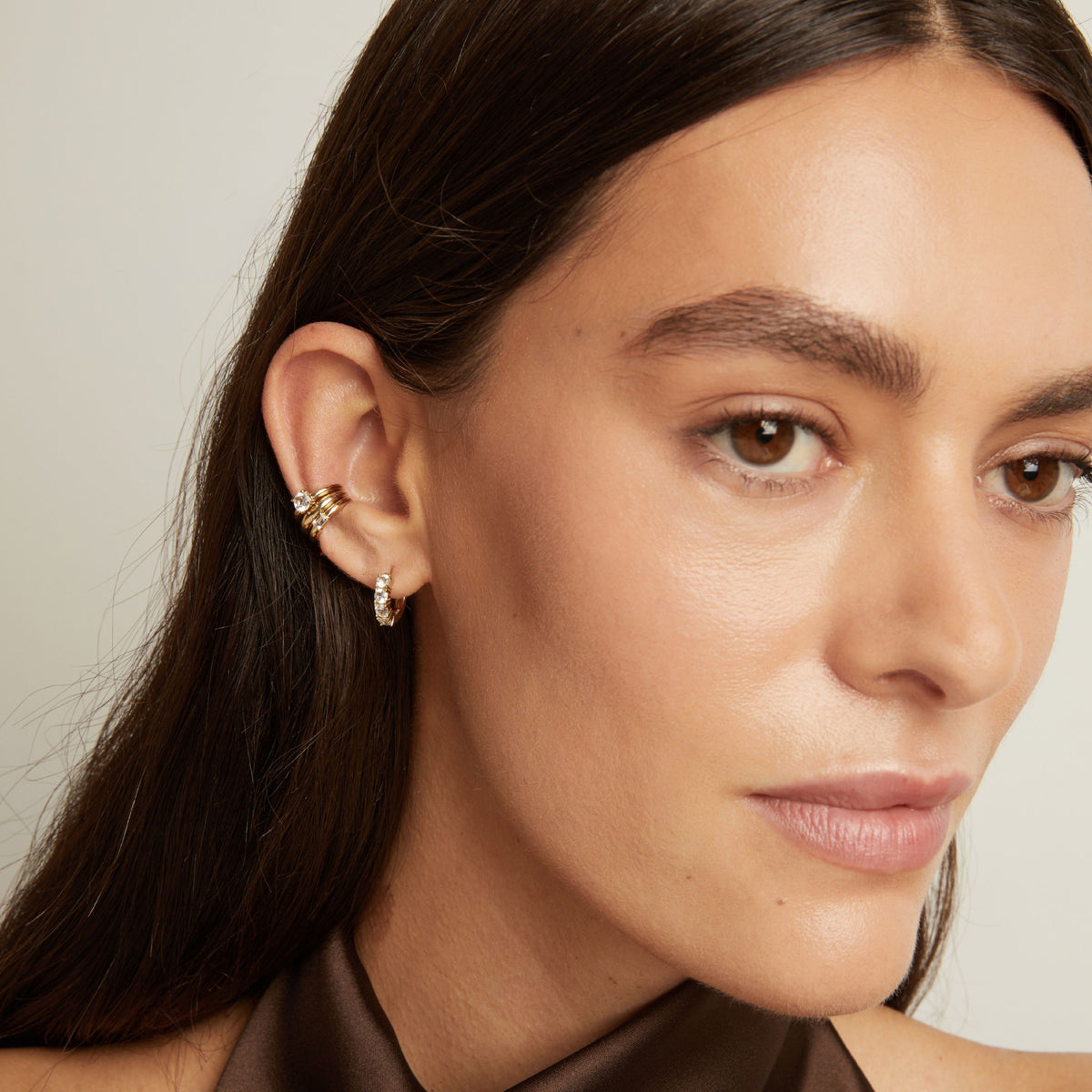 Model. Close up of woman wearing three gold ear cuffs and a huggie earring with white sapphires on neutral background. 