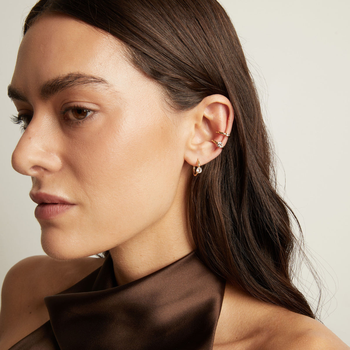 Model. Close up of woman wearing two gold ear cuffs and a huggie earring with white sapphires on neutral background. 