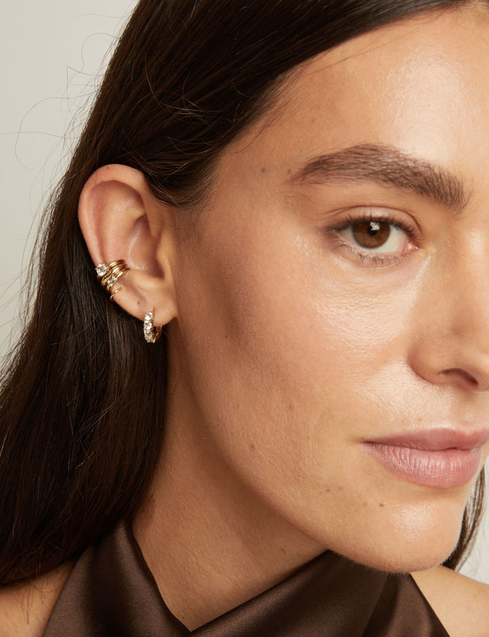 Model. Close up of woman wearing three gold ear cuffs and a huggie earring with white sapphires on neutral background. 