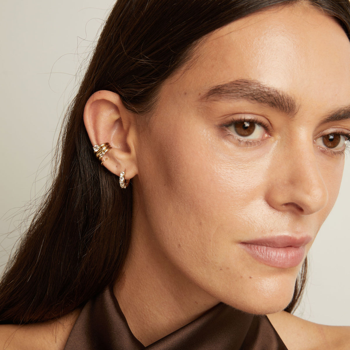 Model. Close up of woman wearing three gold ear cuffs and a huggie earring with white sapphires on neutral background. 