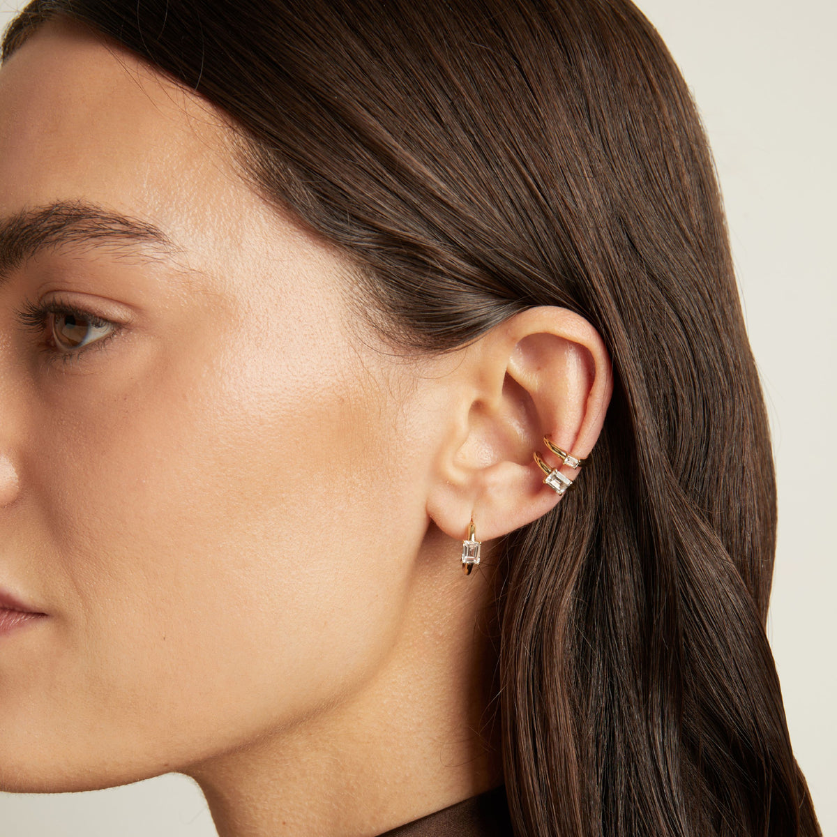 Model. Close up of woman wearing two gold ear cuffs and a huggie earring with white sapphires on neutral background. 
