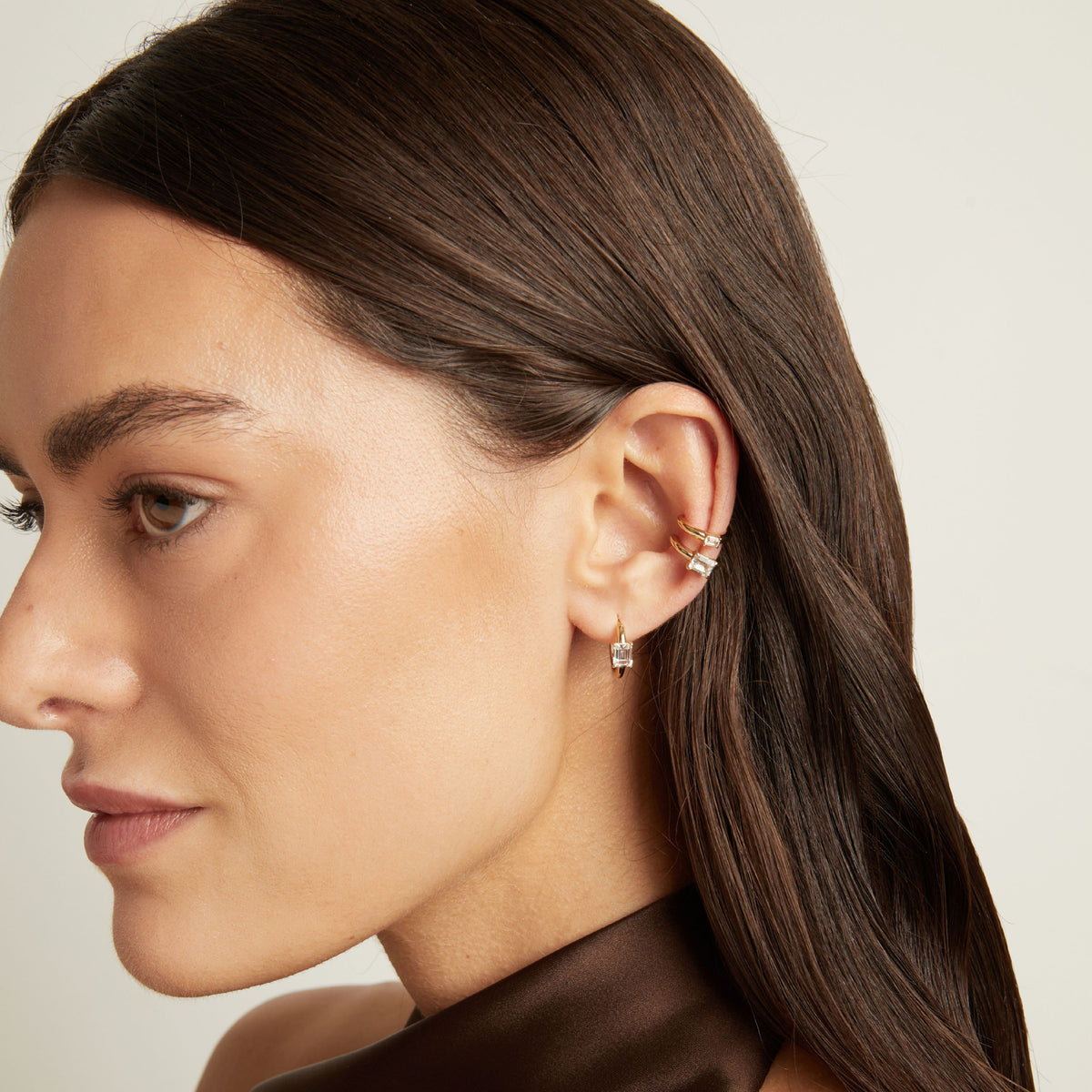 Model. Close up of woman wearing two gold ear cuffs and a huggie earring with white sapphires on neutral background. 