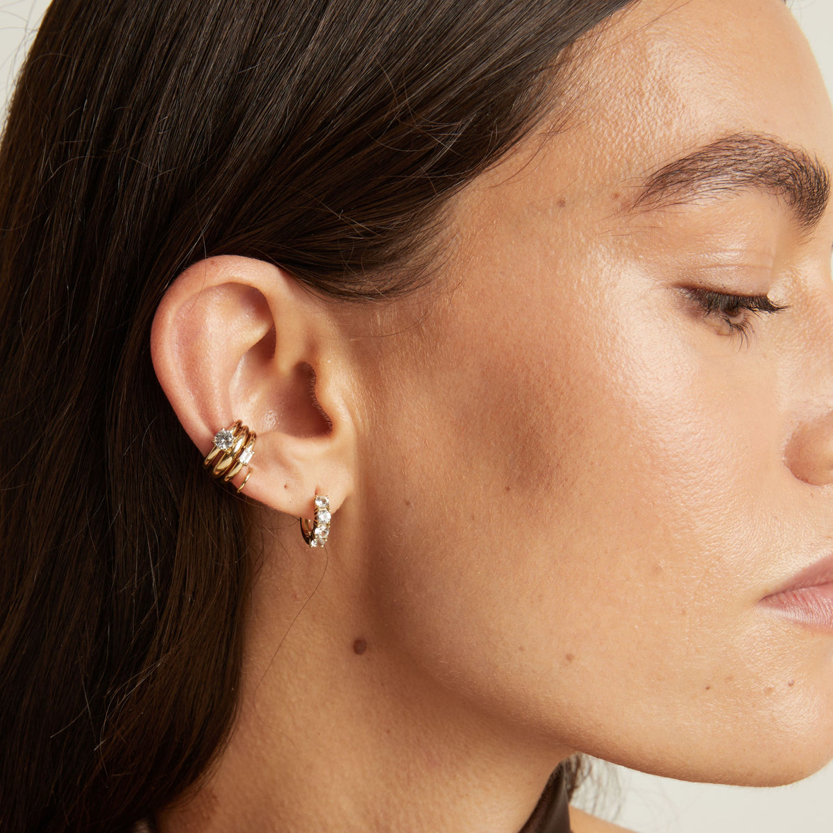 Close up of woman wearing three gold ear cuffs and a huggie earring with white sapphires on neutral background. 