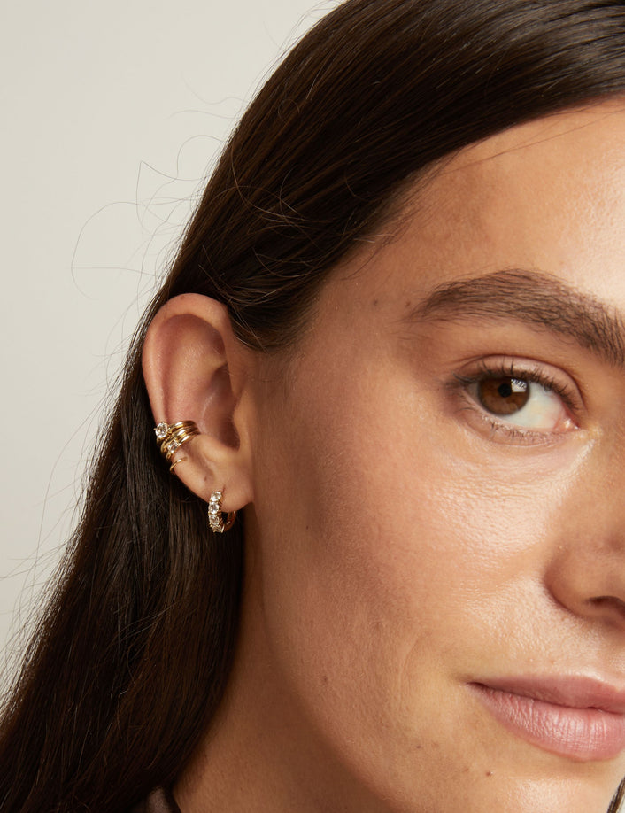 Close up of woman wearing three gold ear cuffs and a huggie earring with white sapphires on neutral background. 