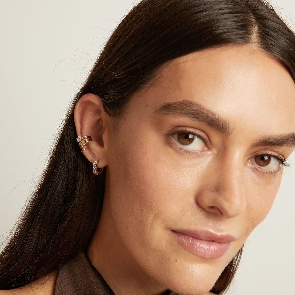 Close up of woman wearing three gold ear cuffs and a huggie earring with white sapphires on neutral background. 