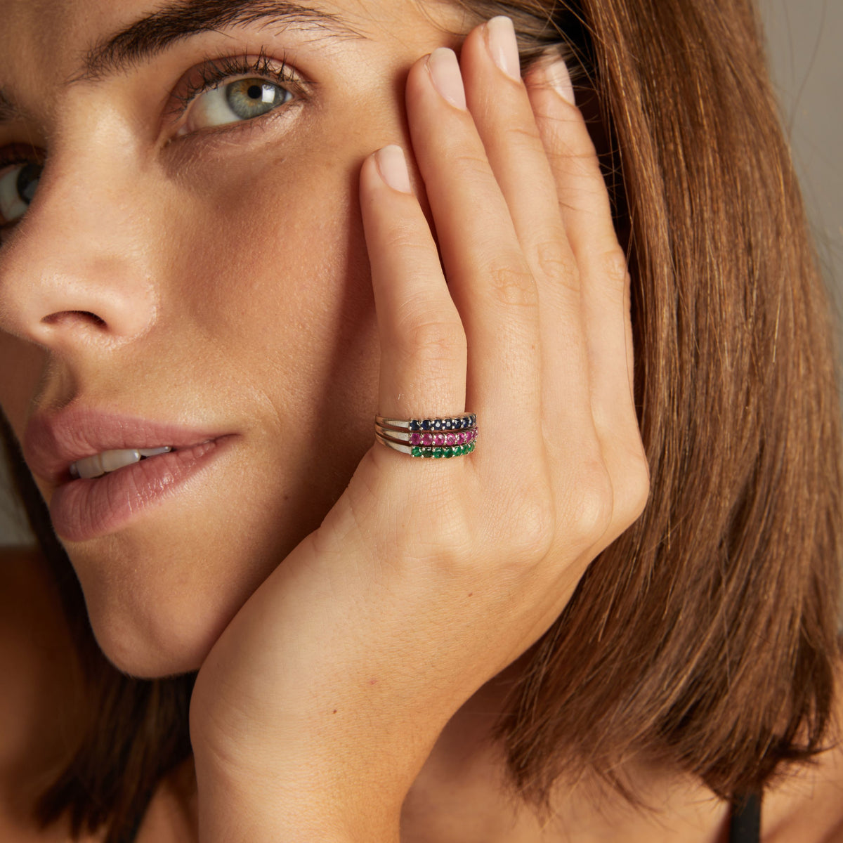 Model. Close up of woman wearing a ring with rows of blue sapphire, red ruby and green emerald gemstones on neutral background.
