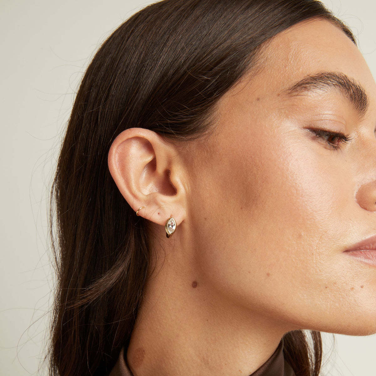 Close up of woman wearing a gold bezel huggie earring on neutral background.