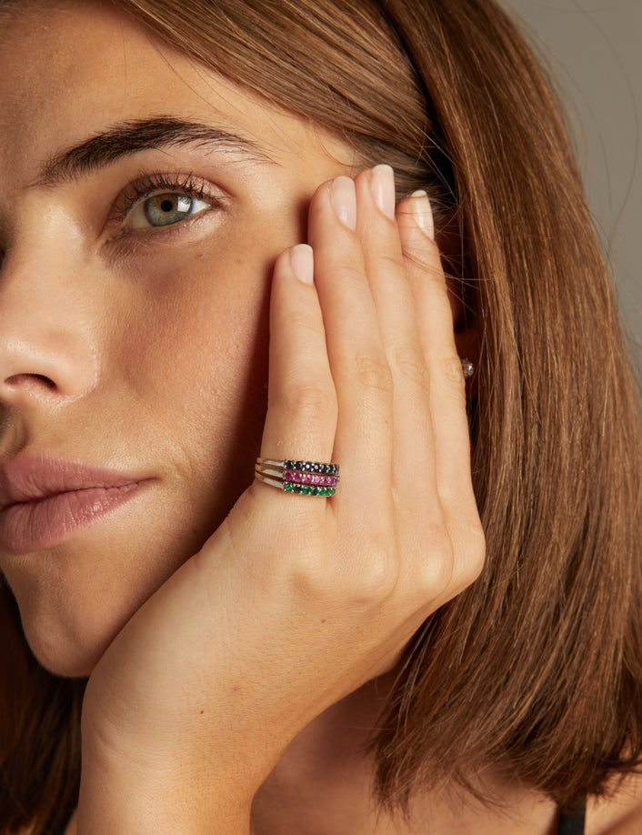 Model. Close up of woman wearing a ring with rows of blue sapphire, red ruby and green emerald gemstones on neutral background.