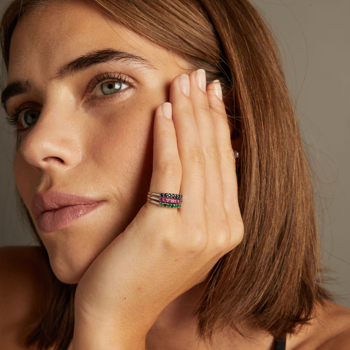 Model. Close up of woman wearing a ring with rows of blue sapphire, red ruby and green emerald gemstones on neutral background.