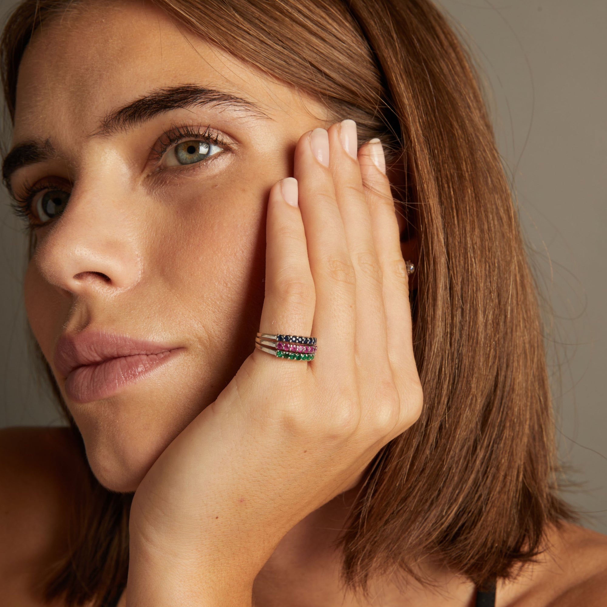 Model. Close up of woman wearing a ring with rows of blue sapphire, red ruby and green emerald gemstones on neutral background.
