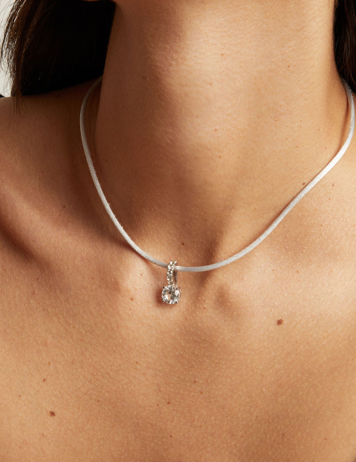 Model. A close-up of a model wearing a thin grey cord necklace with a gemstone pendant, paired with a black camisole top against a light neutral background.