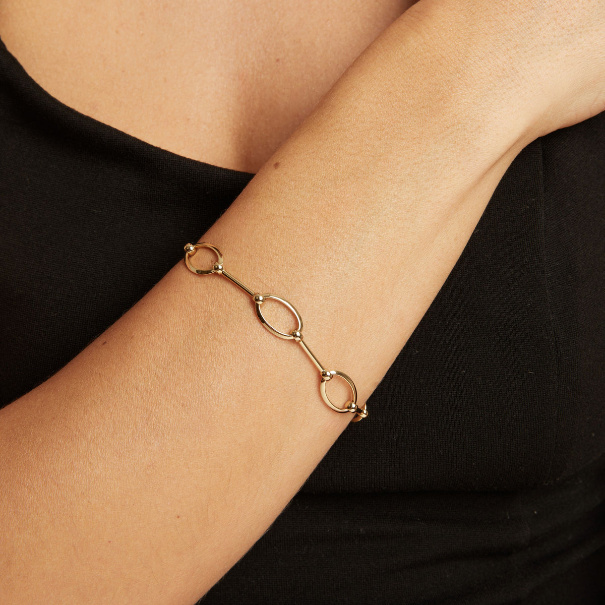 Model. A gold chain bracelet with oval links worn on a woman's wrist, shown against a black top.