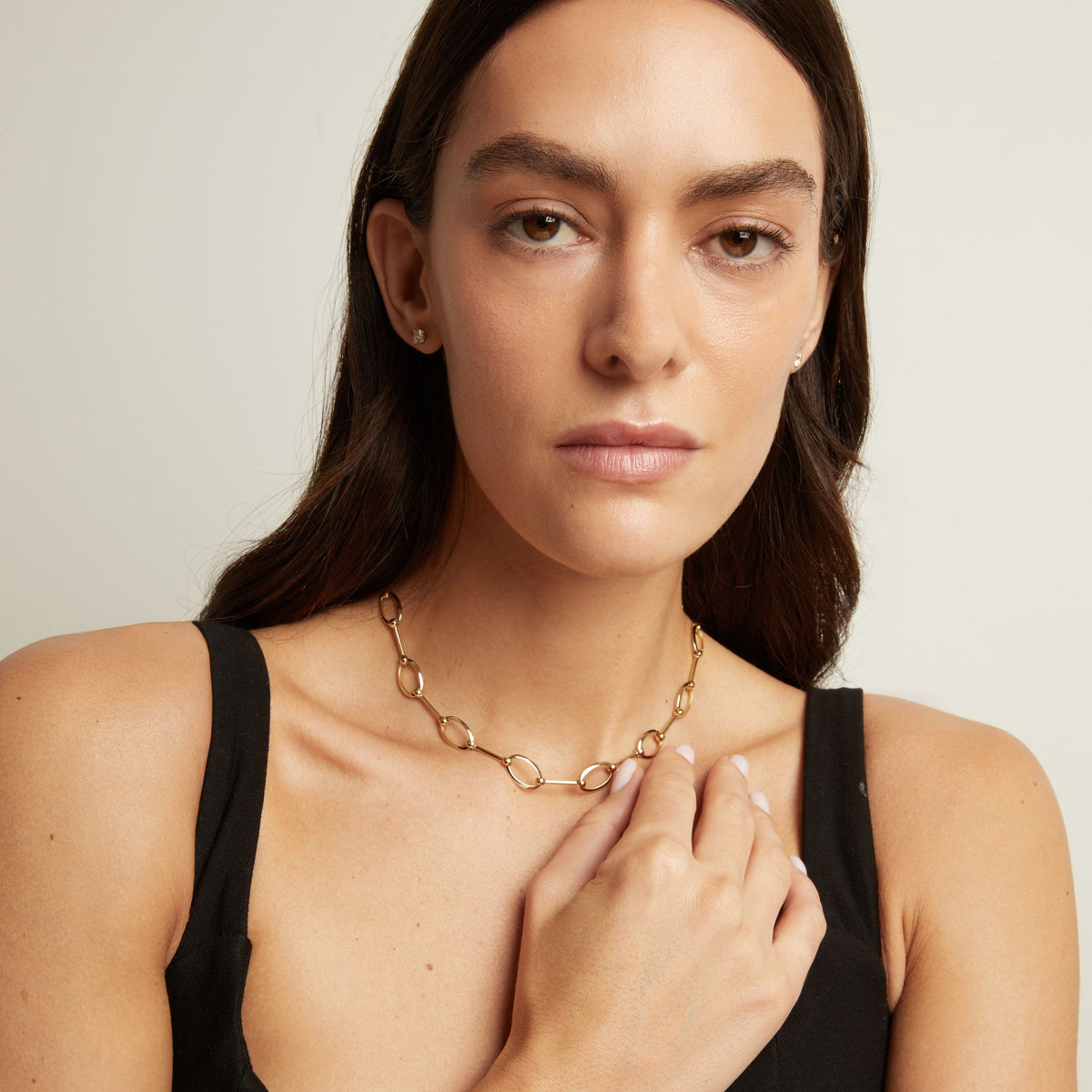 Model. A close-up of a woman wearing a gold chain necklace with interlocking oval links, paired with a black tank top against a light gray background.