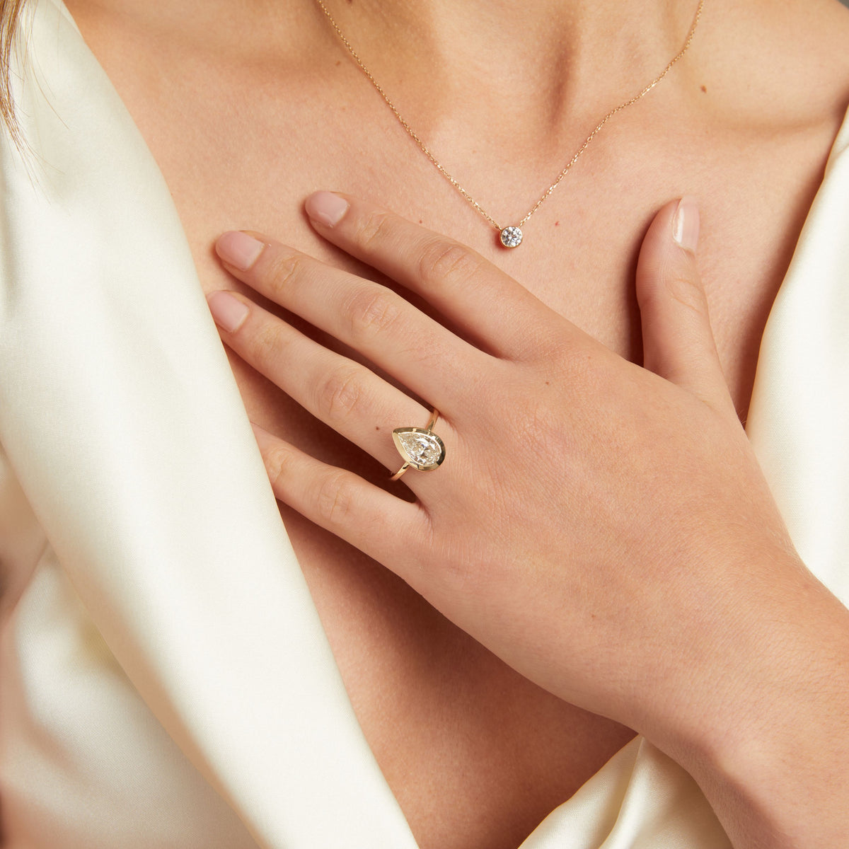 Model. A close-up lifestyle shot of a woman's hand wearing a gold ring with a pear bezel-set diamond. She is wearing a cream satin top against a neutral gray background.