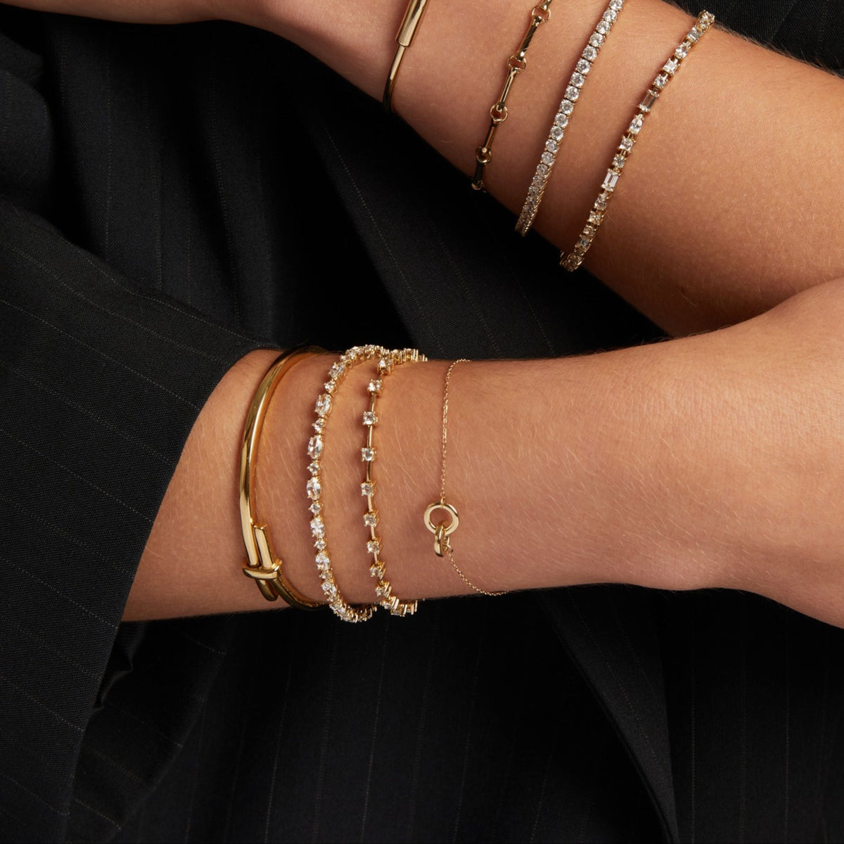 Model. A close-up of a person's wrists wearing multiple delicate gold bracelets and bangles, styled against a black pinstripe blazer.