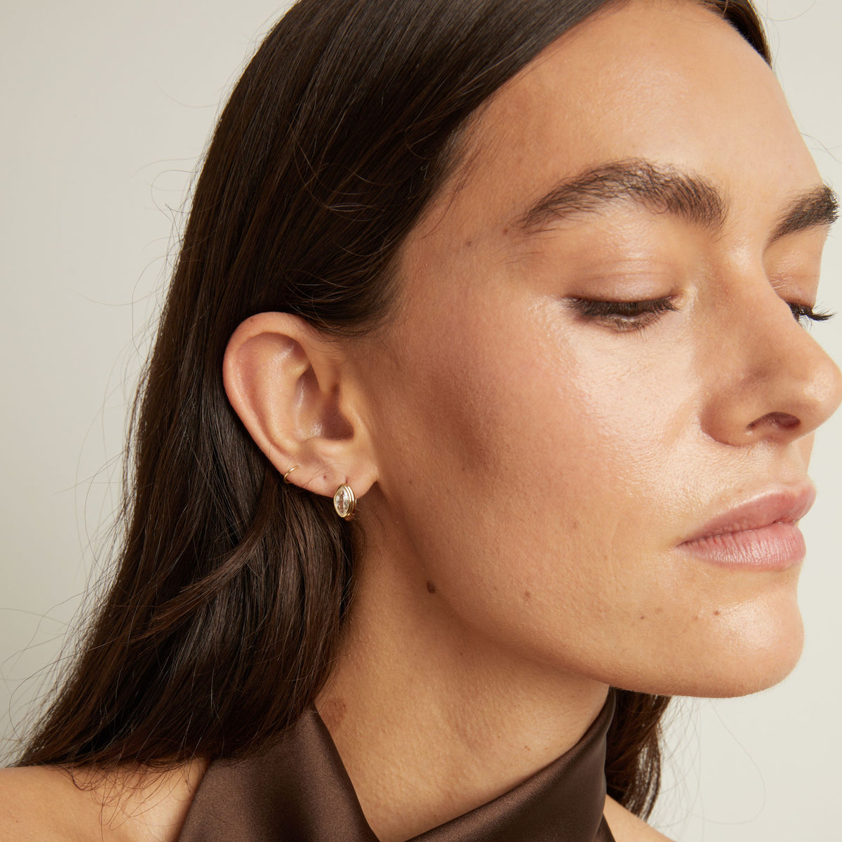 Close up of woman wearing a gold bezel huggie earring on neutral background.