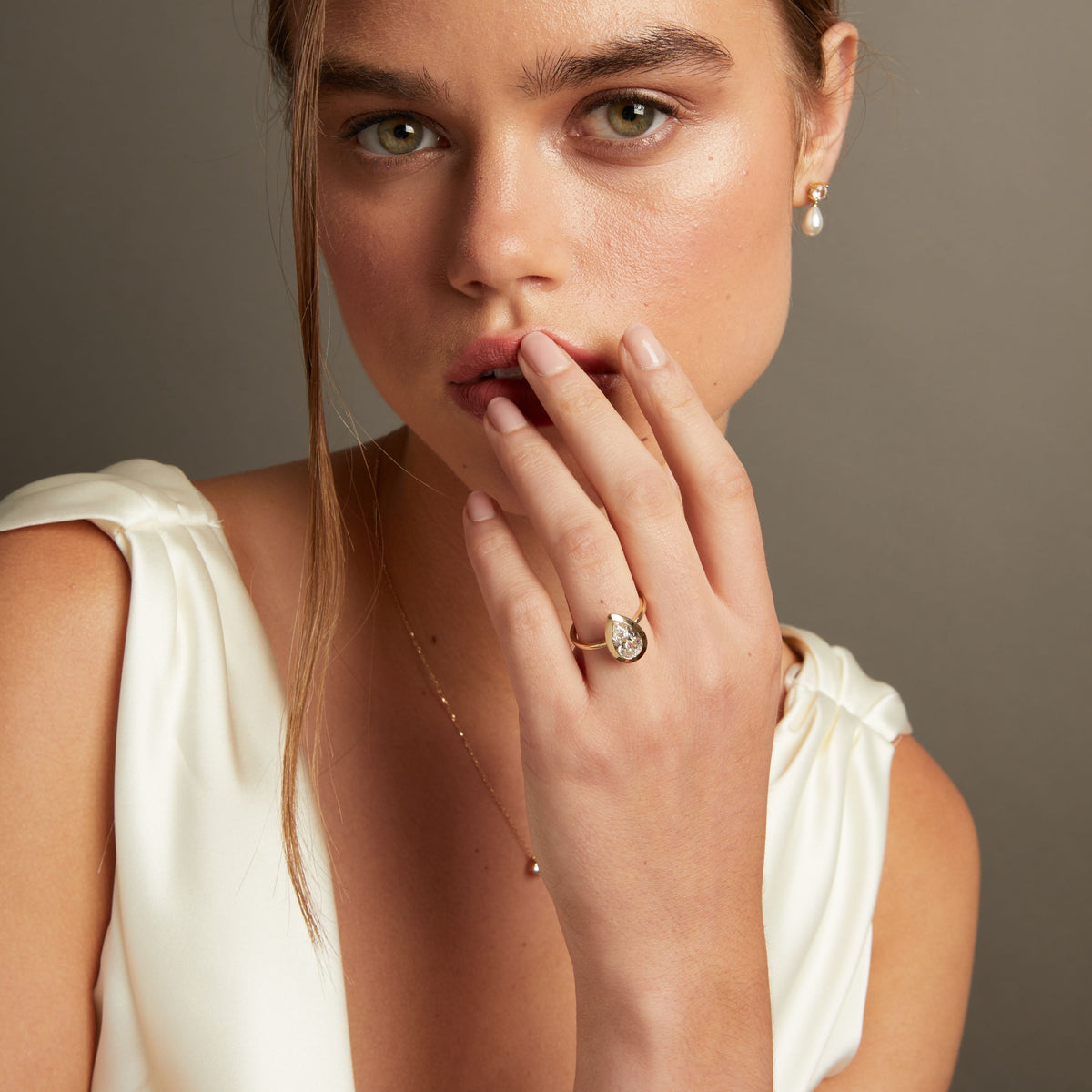 Model. A close-up lifestyle shot of a woman's hand wearing a gold ring with a pear bezel-set diamond. She is wearing a cream satin top against a neutral gray background.