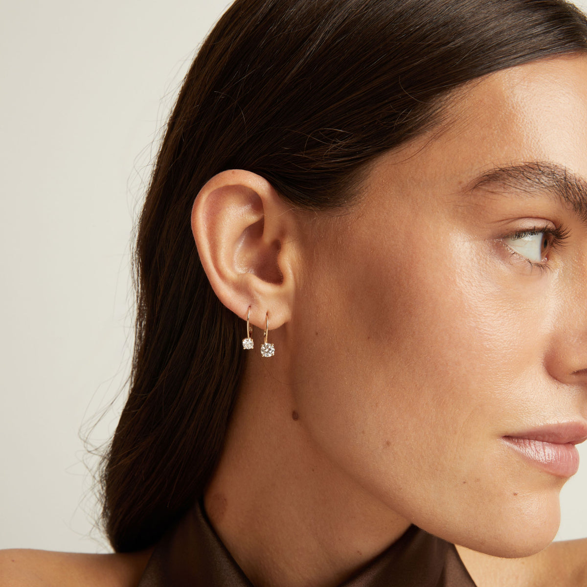 Model. Close up of a woman wearing 2 gold hoop earrings with diamonds on neutral background.
