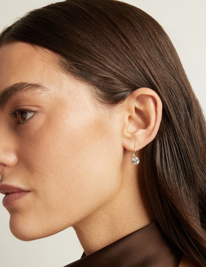 Close up of a woman wearing gold hoop earring with white gemstone on neutral background.