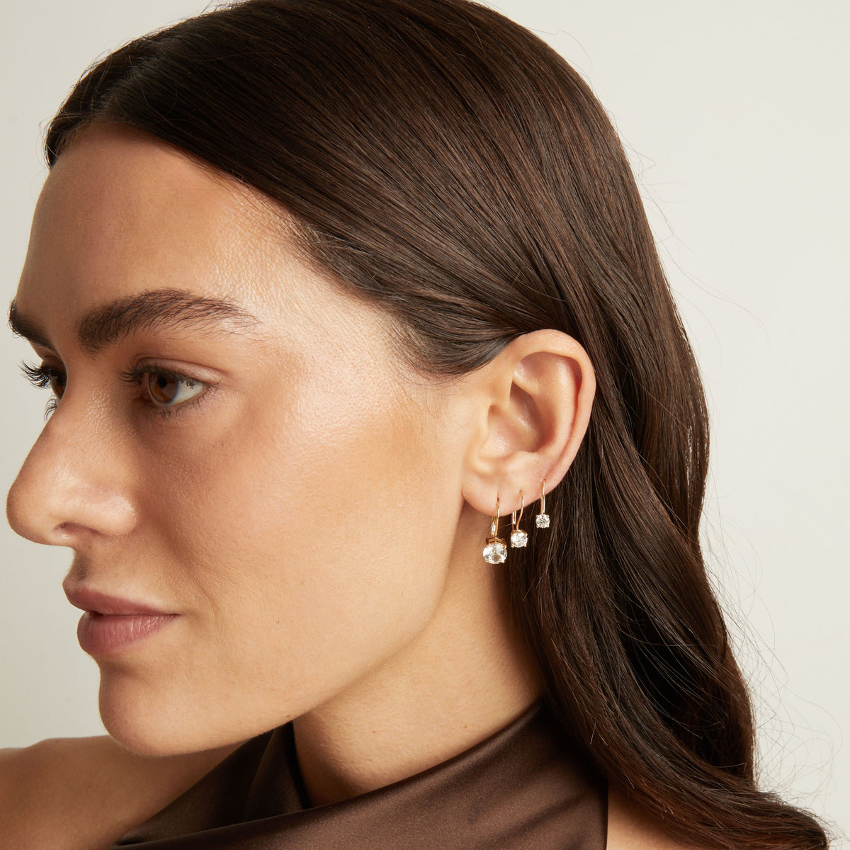 Model. Close up of a woman wearing 3 gold hoop earrings with white gemstones on neutral background.