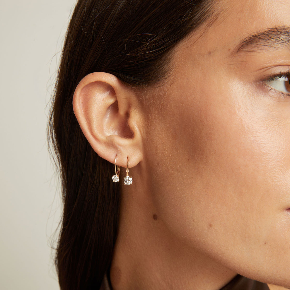 Model. Close up of a woman wearing 2 gold hoop earrings with diamonds on neutral background.
