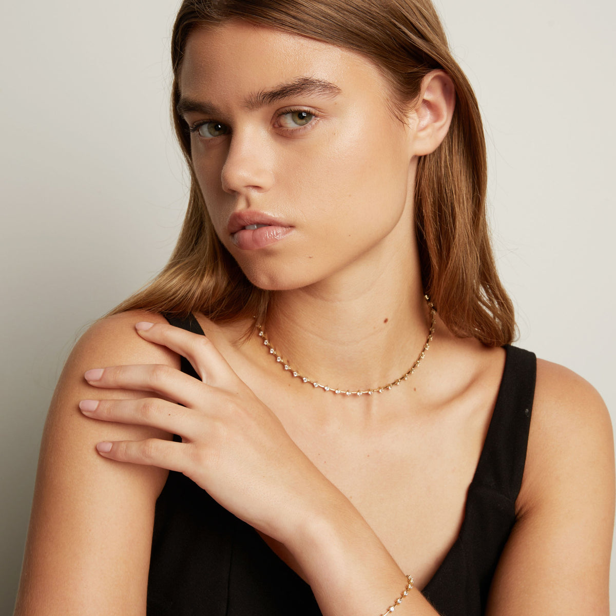 Model. A close-up of a model wearing a delicate gold chain necklace with small clear stones, set against a soft neutral background.