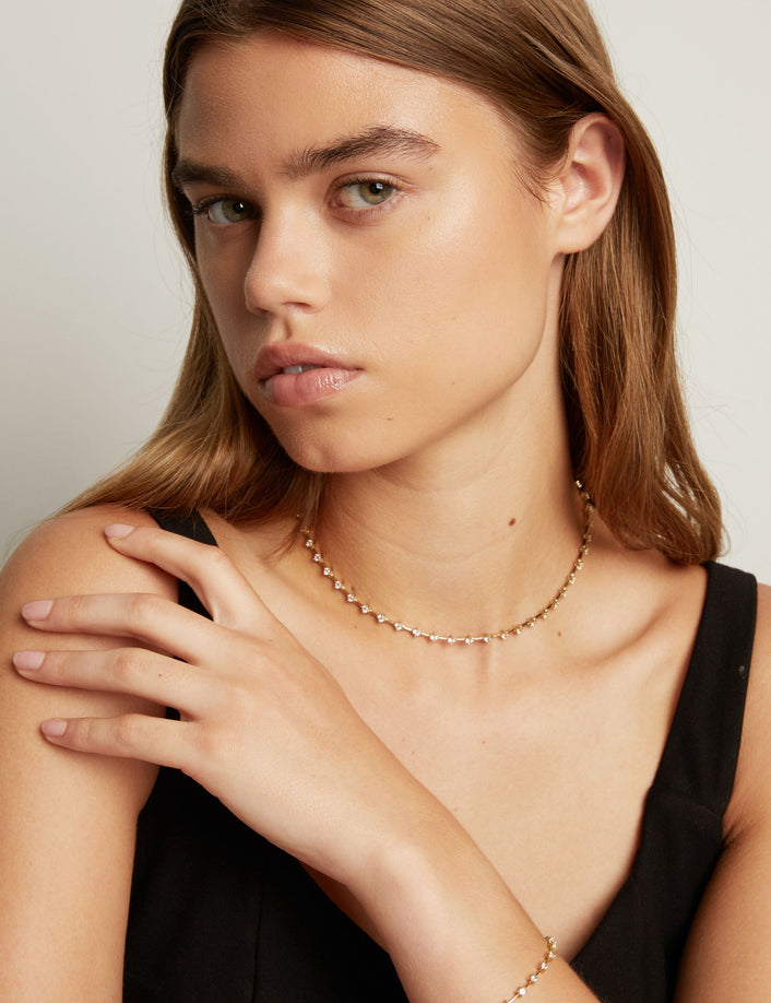 A close-up of a model wearing a delicate gold chain necklace with small clear stones, set against a soft neutral background.