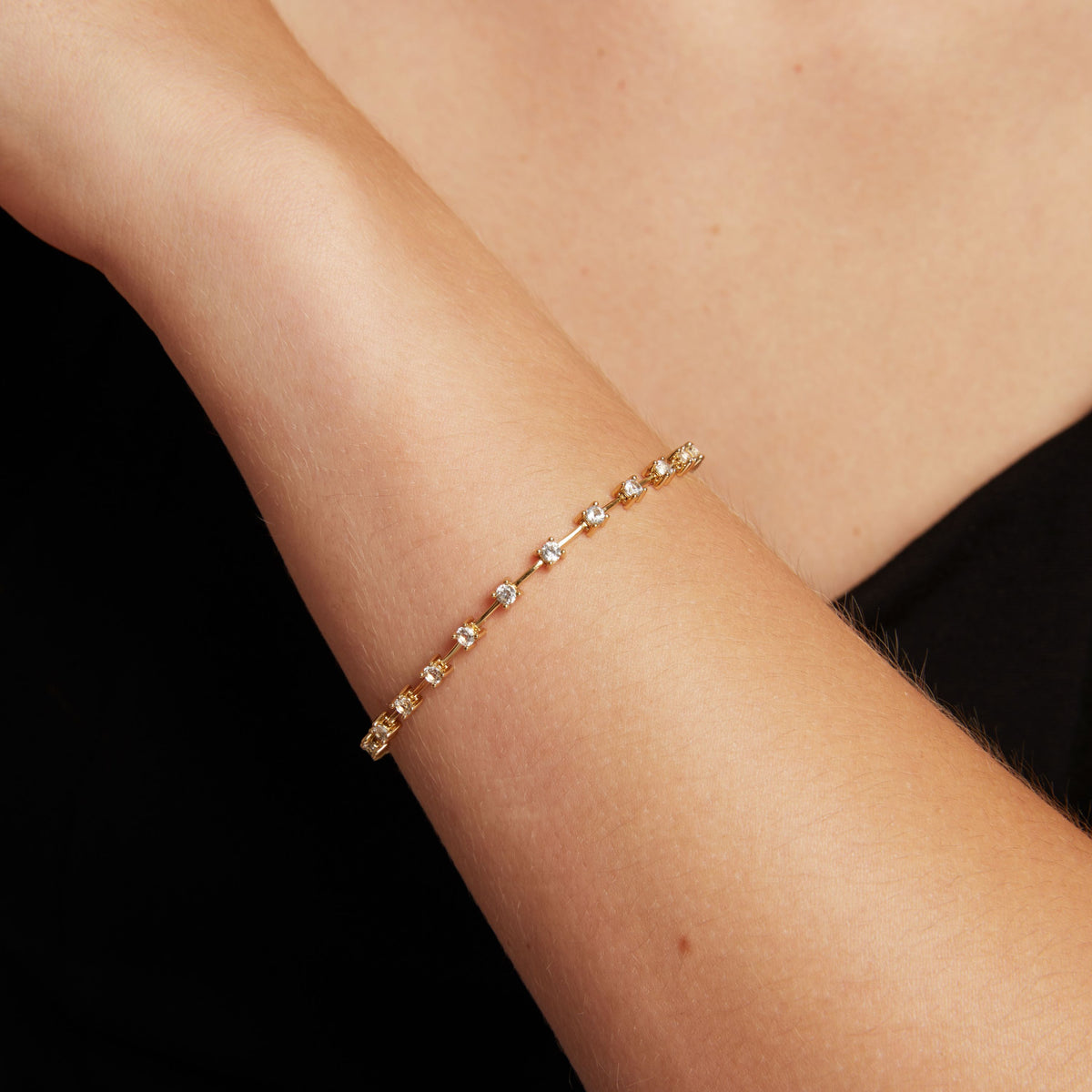 Model. A delicate gold bracelet with small clear gemstones, shown worn on a person's wrist against a dark background.