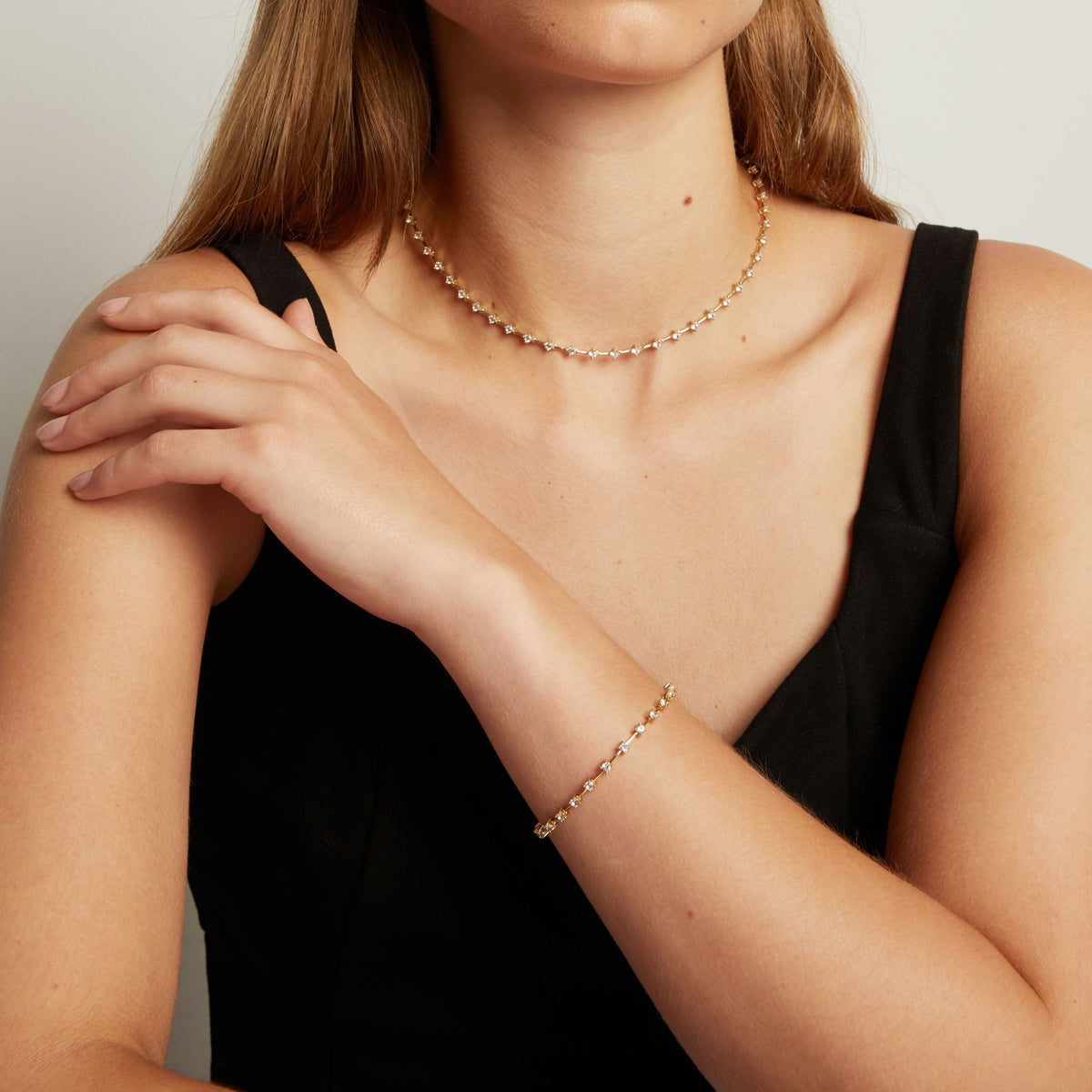 Model. A close-up of a model wearing a delicate gold chain necklace and a matching gold beaded bracelet, paired with a black sleeveless top against a neutral light gray background.
