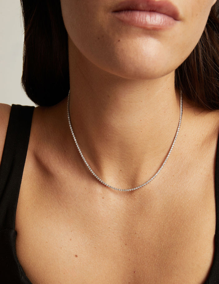 A close-up of a delicate silver tennis necklace worn by a woman, showing the fine chain draped around the neck over a black top.