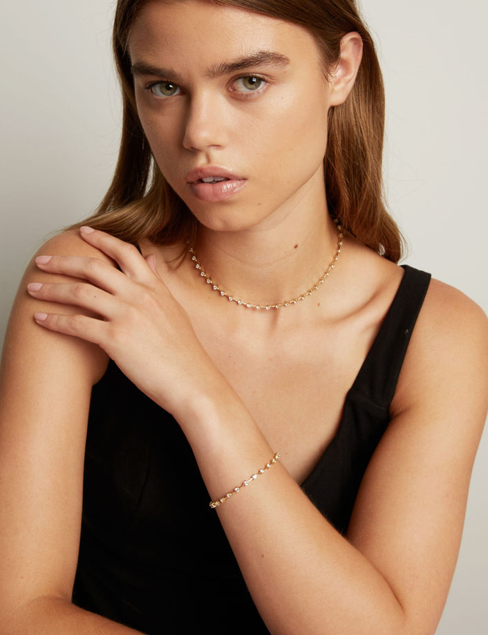 A close-up of a model wearing a delicate gold chain necklace and a matching gold beaded bracelet, paired with a black sleeveless top against a neutral light gray background.