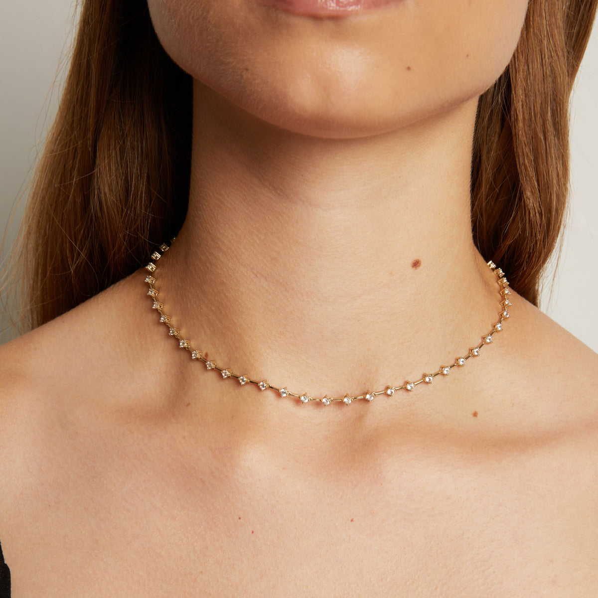 Model. A close-up of a model's neck and upper chest wearing a delicate gold chain necklace with small clear stones, set against a soft neutral background.