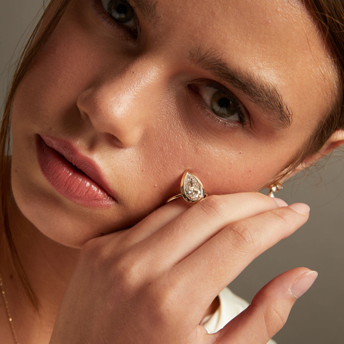 Model. A close-up lifestyle shot of a model resting a gold ring with a pear-shaped diamond on her hand and face.