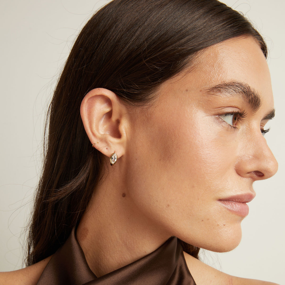 Model. Close up of woman wearing a gold bezel huggie earring on neutral background.