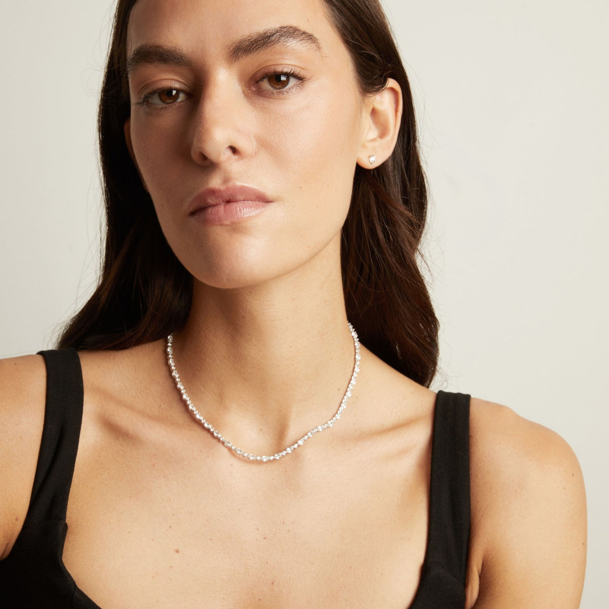 Model. A close-up of a woman wearing a delicate, tennis necklace with a matching white stud earring, styled with a black tank top against a light gray background.