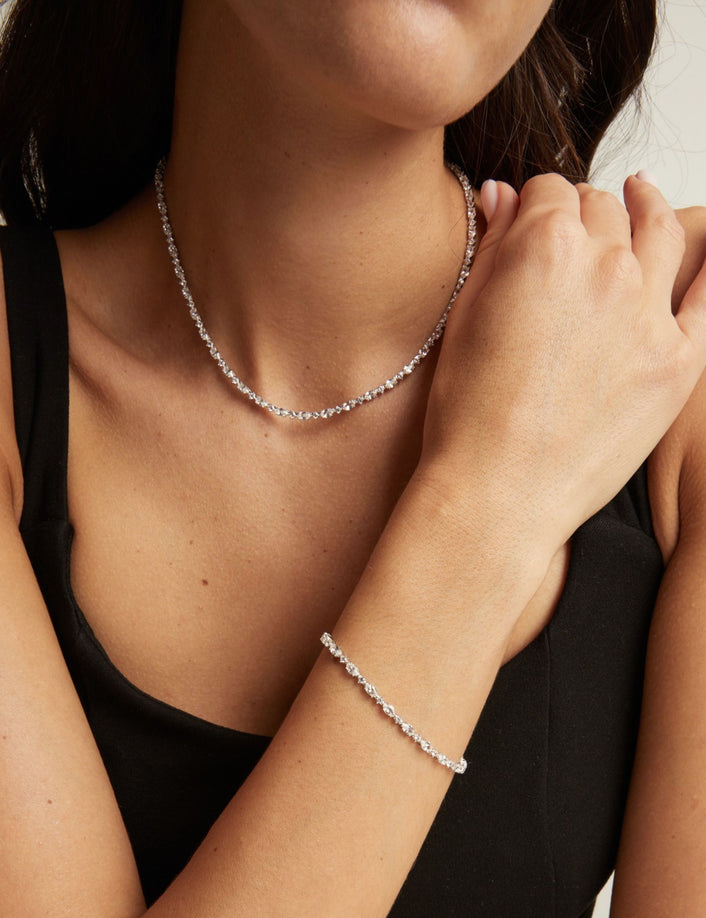 Model. A close-up of a woman wearing a delicate silver tennis necklace and a matching silver bracelet, paired with a black sleeveless top against a neutral light gray background.