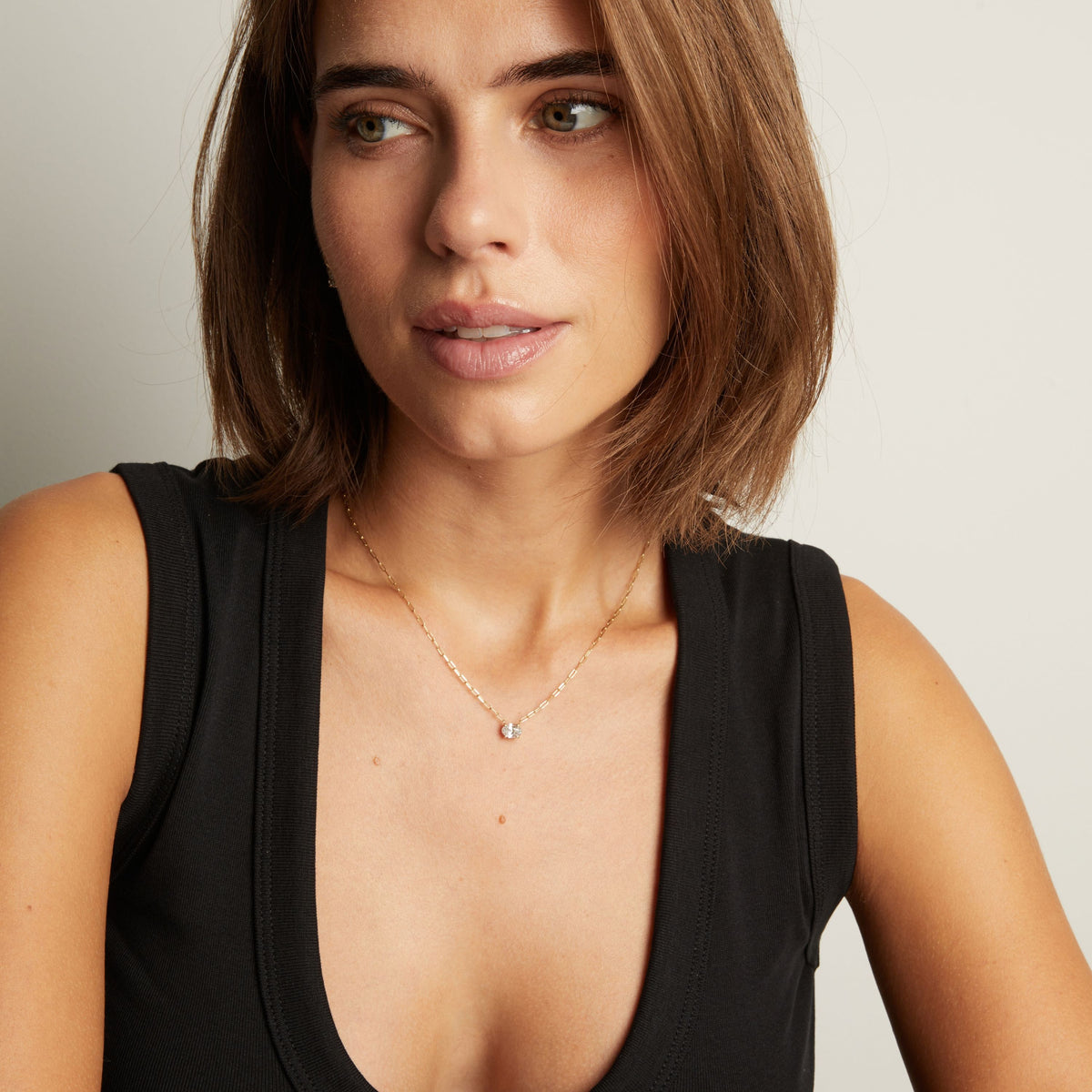 Close up of a woman in a black top wearing a chain necklace with a solitaire oval diamond on neutral background.