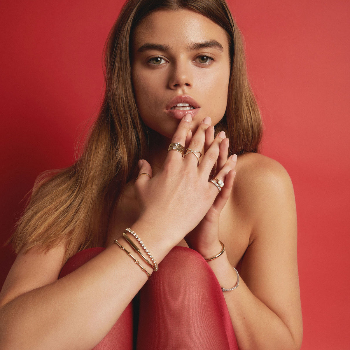 Model. A woman with long light brown hair poses against a solid red background, wearing multiple delicate gold rings and stacked gold and silver bracelets on her hands.