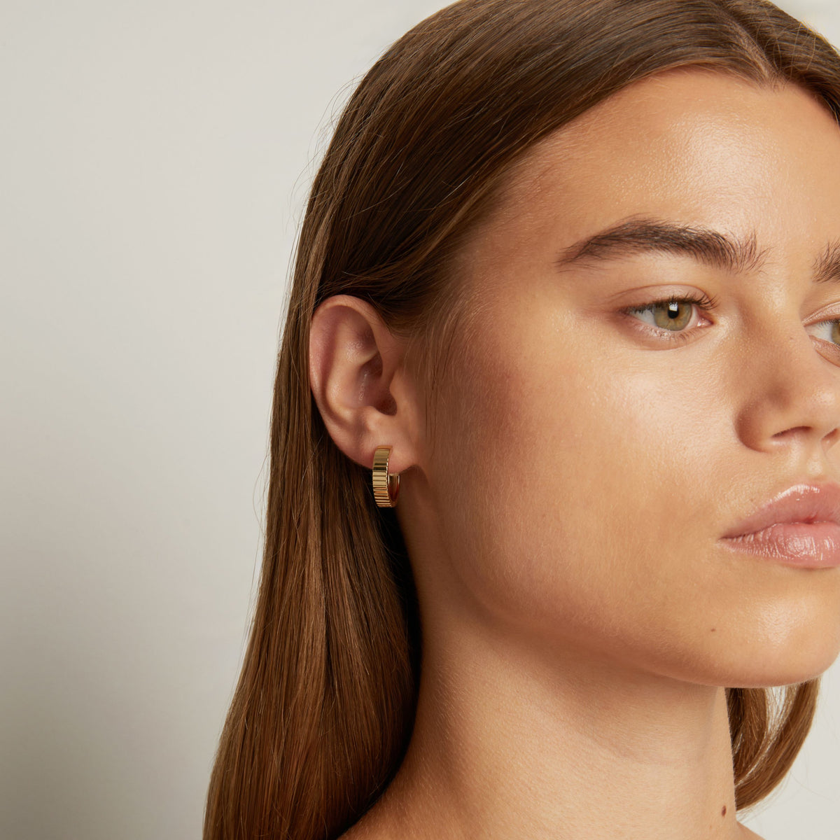 Model. Close up shot of woman with long brown hair wearing a chunky gold hoop earring in neutral background.