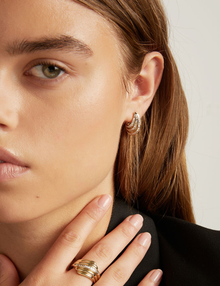 A close-up of a woman with light brown hair wearing a small gold hoop earring with textured, baguette gemstone detailing, set against a plain light gray background.